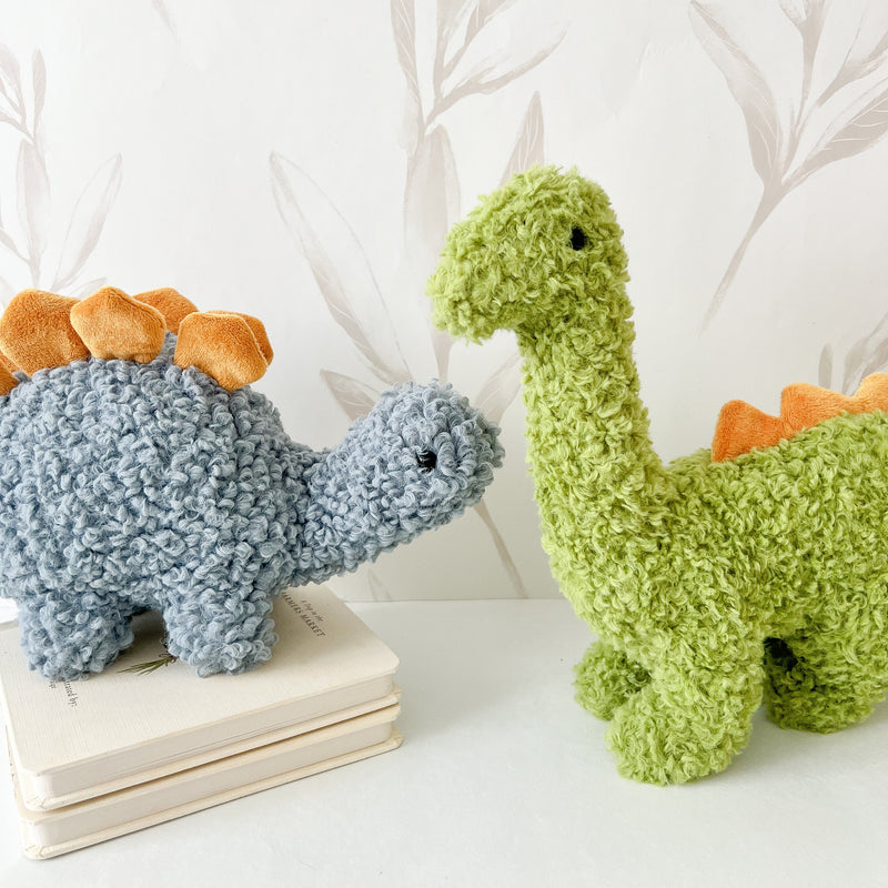 Two plush dinosaur toys, one blue and one green, on a white surface with a white decorative branch background.