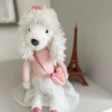 White dog toy with a pink scarf and dress on a shelf with a small model of the Eiffel Tower in the background.